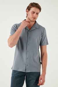 Camisa gris de manga corta con botones y cuello de punta, con un tejido texturizado y cinco botones en la parte frontal, combinada con jeans azules.