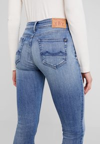 Blå denimjeans med en åtsittande design, med subtil blekning, två bakfickor och en beige läderlogg på midjan.