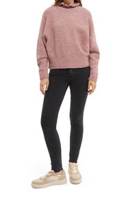 Suéter de punto rosa con un escote con volantes, combinado con jeans negros ajustados y zapatillas beige con detalles en rosa. Apariencia suave y texturizada.