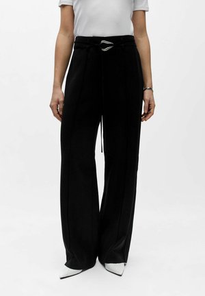 Žena v čiernych širokých vysokopríponových nohaviciach s čiernym opaskom a bielymi špicatými topánkami, stojaca pred jednofarebným pozadím.