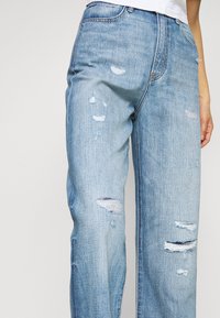 Jeans de denim azul claro con un corte relajado, detalles desgastados y costuras visibles. Con desgarros notables y bordes deshilachados por todo el pantalón.