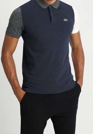 Mann trägt ein kurzärmeliges marineblaues Poloshirt mit grauen und weißen Ärmeln und schwarze Hose, Hände in den Taschen, steht vor einem einfachen Hintergrund.