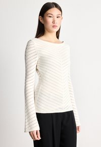 Strikket topp i off-white med lange ermer, med chevronmønstrede utskjæringer og vid hals, kombinert med svarte bukser.