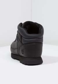 Botín de cuero negro hasta el tobillo con cuello acolchado, puntera redonda y suela de goma resistente. Presenta un detalle de logo en el lado externo y un diseño de cordones.