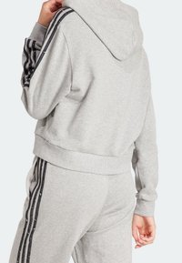 Sudadera con capucha gris con detalles de rayas negras en las mangas, hecha de material suave, con un corte relajado y puños y dobladillo acanalados.