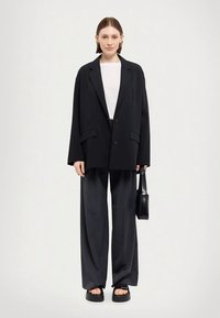 Ylisuuri musta bleiseri valkoisen paidan päällä, yhdistetty leveisiin harmaisiin housuihin ja mustiin korollisiin sandaaleihin, kädessä pieni musta laukku.