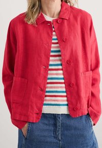 Woman wearing a red buttoned jacket over a striped sweater with white, blue, pink, and red stripes, paired with blue denim pants.