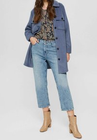 Cappotto di lana azzurro chiaro con chiusura a bottoni e due tasche sul petto, abbinato a un top scuro con fantasia e jeans a gamba dritta in denim azzurro chiaro strappati. Stivali alla caviglia beige.