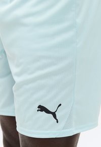 Φωτεινά μπλε αθλητικά σορτς σε άτομο, με μαύρο λογότυπο Puma κοντά στο κάτω μέρος στο αριστερό πόδι.