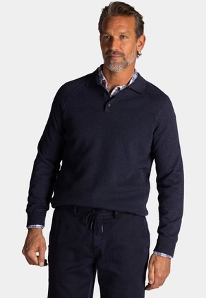 Man met kort haar en baard, gekleed in een marineblauwe trui over een geruit overhemd en donkere broek, staand tegen een effen achtergrond.