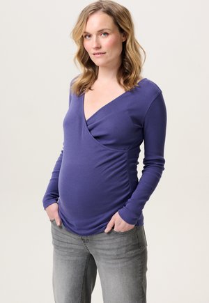 Lila langärmliges Schwangerschaftsoberteil mit V-Ausschnitt und Raffung an der Taille, kombiniert mit grauen Jeans für einen lässigen Look.