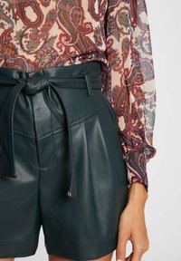 Shorts en similicuir vert foncé avec une taille ceinturée, des plis et une texture brillante, associés à un chemisier clair à motifs.