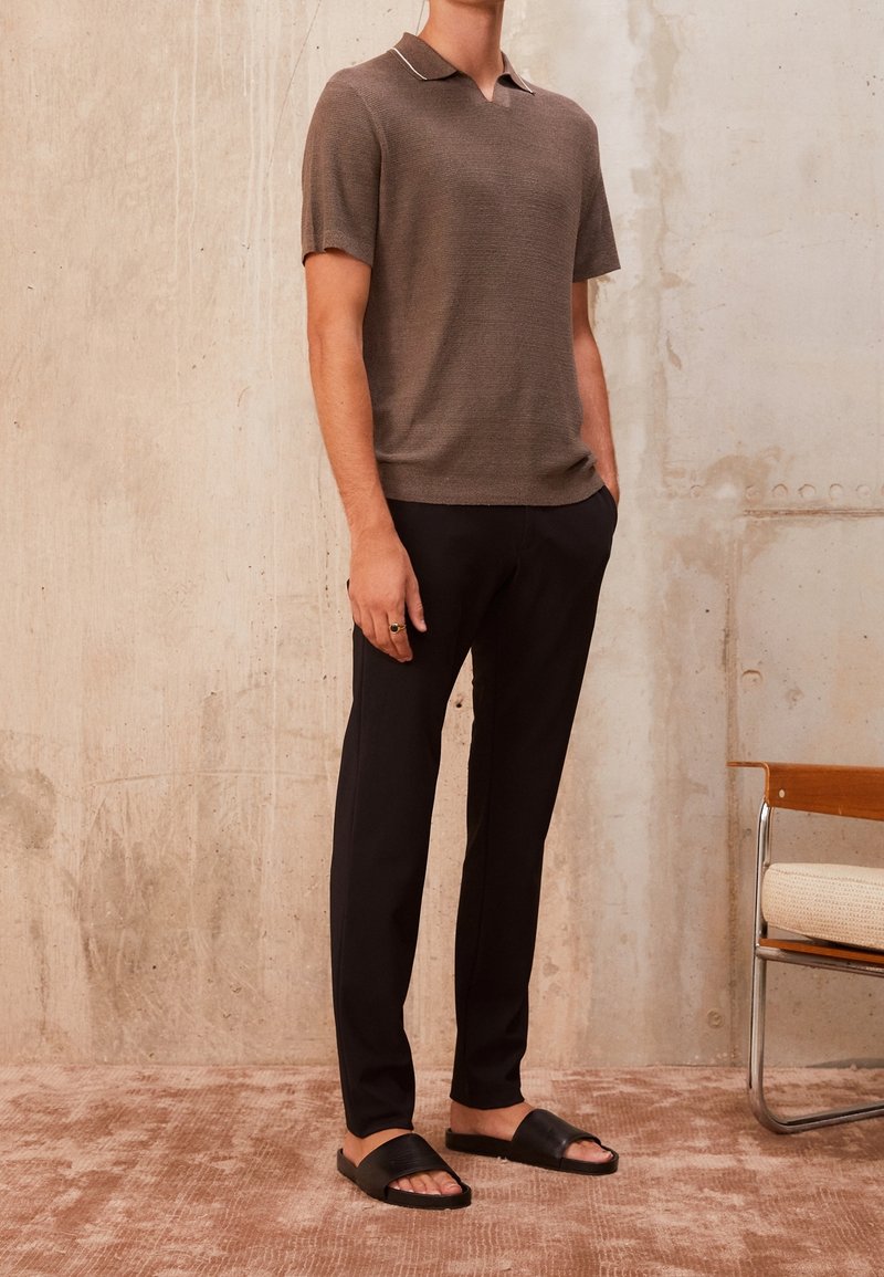 Homme portant un polo en maille marron à manches courtes, pantalon noir fuselé, sandales noires à enfiler, debout contre un mur beige texturé près d'une chaise en bois.