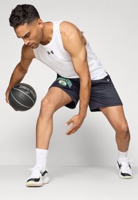 Sportstøj bestående af en hvid tanktop og mørke shorts med "BOSTON" grafik. Holder en sort basketball, iført hvide og sorte sneakers.