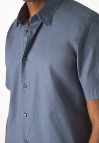 Kortärmad skjorta i mörkblått tyg, med spetsiga kragar, sex svarta knappar och en slät yta.