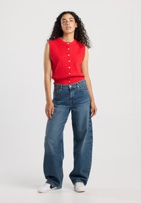 Red sleeveless knit top with a round neckline and button detail, paired with loose-fitting blue denim jeans and white sneakers.