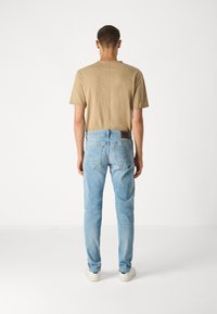 Moški stoji obrnjeno stran od kamere, oblečen v bež majico, svetlo modre kavbojke z zadnjimi žepi in bele športne čevlje na beli podlagi.