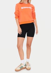 Orange sweatshirt med "BISOU" text, svarta cykelbyxor och beige sneakers med gröna ränder. Vita crew-strumpor. Släta texturer.