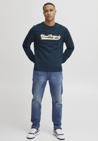 Marine sweatshirt met "Blend Original" grafiek in lichtoranje, gecombineerd met blauwe spijkerbroek en zwart-witte sportschoenen, staande pose.
