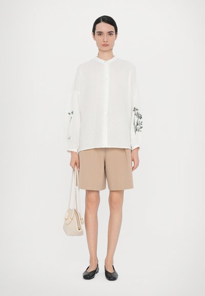 Hvit lin skjorte med knappestolpe og broderte blomsterdesign på ermene, sammen med tan shorts og svarte leiligheter, holder en vevd beige håndveske.