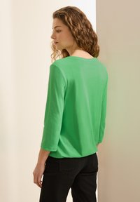Grön, långärmad topp i lätt material. Har en rund halsringning och en avslappnad passform, kombinerad med svarta byxor.