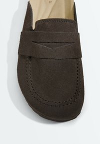 Mocassin en daim marron foncé avec des détails cousus et une large bride sur le dessus avant, vu de haut.