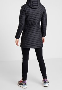 Persoon die een lange zwarte doorgestikte jas draagt, zwarte legging en grijze sportschoenen met paarse en rode details, met de rug naar de kijker toe.