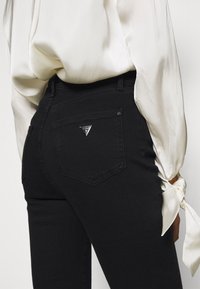 Jeans de mezclilla negra con un diseño ajustado, que presentan un logo triangular en el bolsillo trasero y una textura suave, combinados con una blusa blanca.
