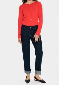 Červený kardigan s knoflíky a dlouhými rukávy, kombinovaný s tmavě modrými džíny s ohrnutými manžetami a černými botami s špičatou špičkou. Hladké textury, jednoduchý design.