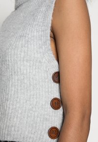 Haut sans manches en maille grise avec une texture côtelée, doté de trois boutons en bois marron sur le côté pour la fermeture et un détail d'accent.