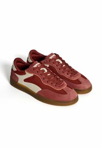 Un par de zapatillas bajas en tonos de rojo y rosa con acentos laterales en beige, suelas de goma marrón y cordones rojos a juego, sobre un fondo blanco.