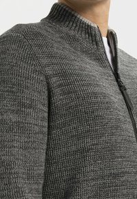 Tummanharmaa vetoketjullinen neulepaita, jossa on teksturoitu neulottu kuvio ja resorikaulus. Varustettu mustalla vetoketjulla ja valkoinen aluspaita näkyy kauluksen alta.