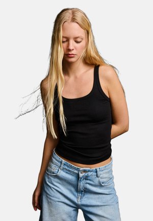 Młoda kobieta z długimi blond włosami, ubrana w czarny top bez rękawów i jasnoniebieskie dżinsy, patrzy w dół, trzymając ręce za plecami.