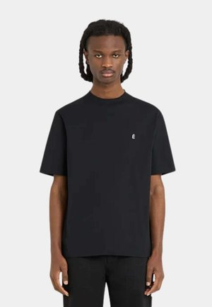Homme aux cheveux tressés portant un t-shirt noir uni à manches courtes avec un petit logo blanc « É » sur la poitrine gauche.