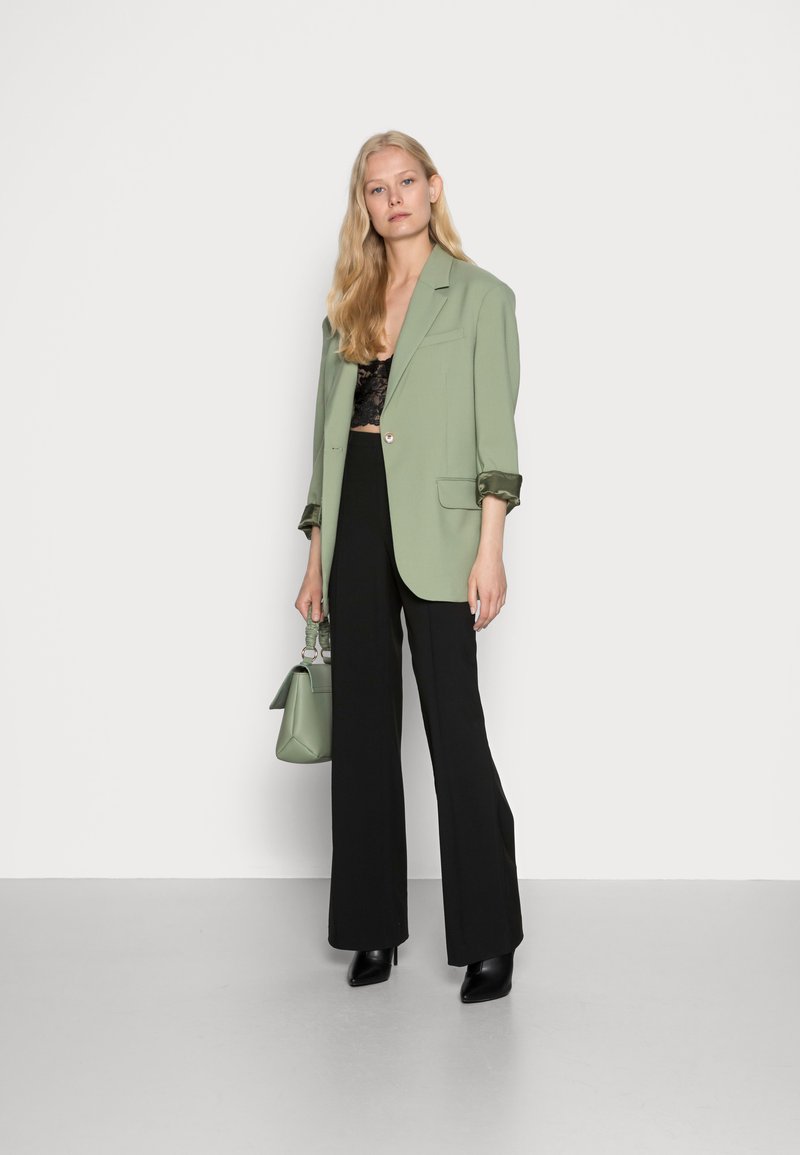 Blazer vert clair avec de grands revers, haut en dentelle noir, pantalon large noir et bottes à talons noires. Portant un petit sac à main vert clair.
