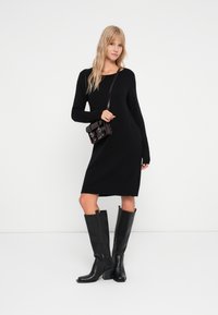 Abito nero in maglia con maniche lunghe e taglio dritto. Abbinato a stivali neri in pelle al ginocchio e una piccola borsa scura.