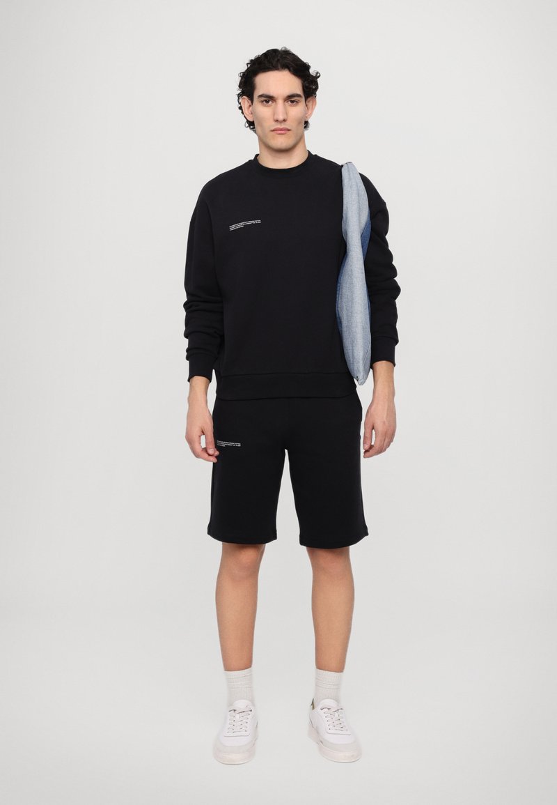 Schwarzer Sweatshirt und schwarze Shorts aus einem Baumwollmix, mit einem grafischen Detail auf der Brust. Weiße Sneakers und graue Socken vervollständigen das Outfit.