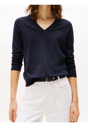 Mujer con suéter azul marino de cuello en V con un pequeño logo en el pecho, pantalones blancos y cinturón negro con hebilla dorada, de pie frente a un fondo blanco.