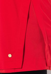 Rode kleding met een gladde textuur, voorzien van een zijsplits en een ronde gouden accenttag aan de linkerzijde.