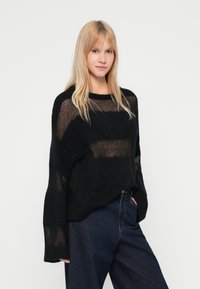 Schwarzer Strickpullover mit einem lockeren, leicht verkürzten Schnitt, der über einen weiten Halsausschnitt und eine weiche, durchsichtige Textur verfügt. Kombiniert mit dunklen Jeans.