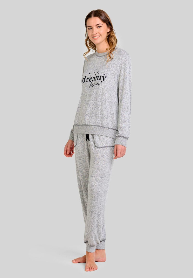 Joven mujer vestida con un conjunto de ropa de lounge gris claro con el texto "soñadora para siempre", de pie descalza frente a un fondo sencillo, sonriendo.