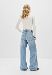 Jean large bleu clair en denim, avec une taille haute, des poches arrière et une texture légèrement délavée. Porté avec un haut à manches longues.