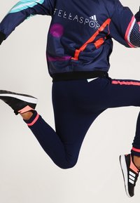 Navy athletic leggings with pink accents, paired with a navy and multicolored jacket featuring geometric patterns and branding. Black athletic shoes.