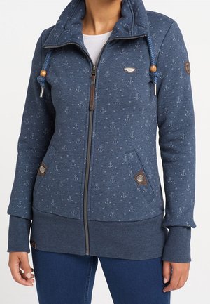 Kobieta ubrana w niebieską bluzę z kapturem zapinaną na suwak, ozdobioną wzorami kotwic i serc, ze sznurkami zakończonymi drewnianymi koralikami oraz kieszeniami z guzikami.