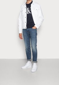 Uomo che indossa una giacca di jeans bianca, una felpa nera con logo, jeans blu risvoltati, calzini bianchi e sneakers bianche, in piedi contro uno sfondo semplice.