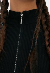Top in tessuto nero a coste con colletto alto e chiusura con zip argentata. Due trecce parzialmente visibili, con perline alle estremità.