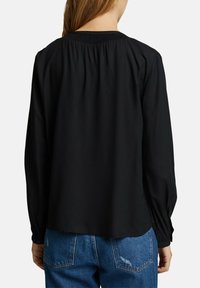 Sort langærmet bluse med samlet rygstykke, blødt materiale og elastiske manchetter, parret med blå denimjeans med baglommer.