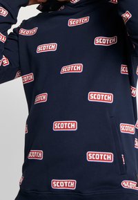 Sudadera azul marino con el logo repetido "SCOTCH" en rojo y blanco. Tejido suave con un bolsillo frontal y capucha ajustable.
