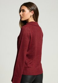 Burgundowa dzianinowa sweter z kołnierzem, ściągaczem u dołu i długim rękawem. Teksturowany materiał o luźnym kroju, z bocznymi rozcięciami na dole.