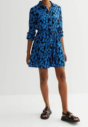 Femme portant une robe à manches longues bleue et noire à motif, avec ceinture à la taille et sandales noires à bouts ouverts, debout sur un sol blanc.